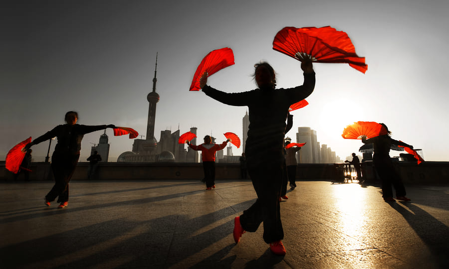 Personas realizando una danza tradicional con abanicos rojos frente al skyline de una ciudad china durante el amanecer.