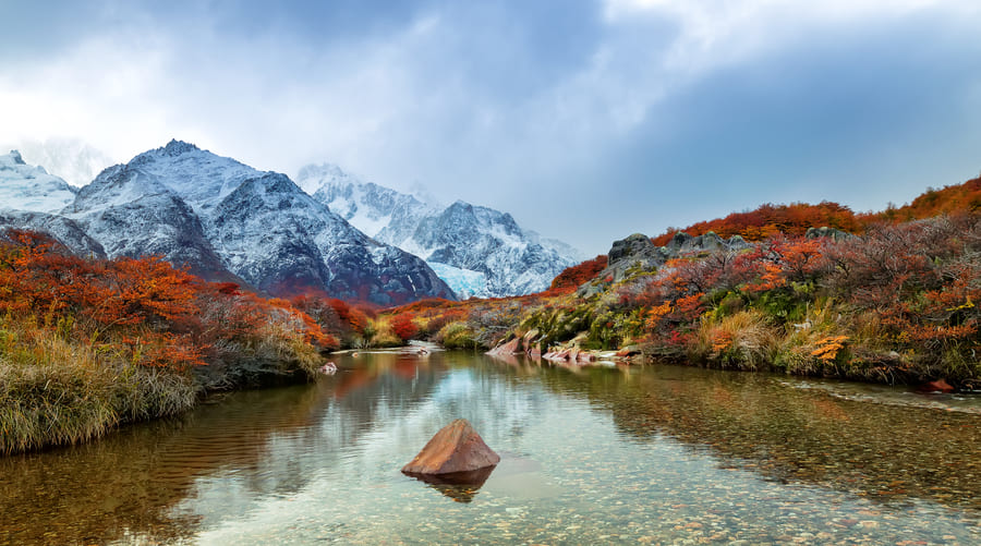 Paisaje natural de China con montañas nevadas, un río de aguas cristalinas y árboles con colores otoñales.