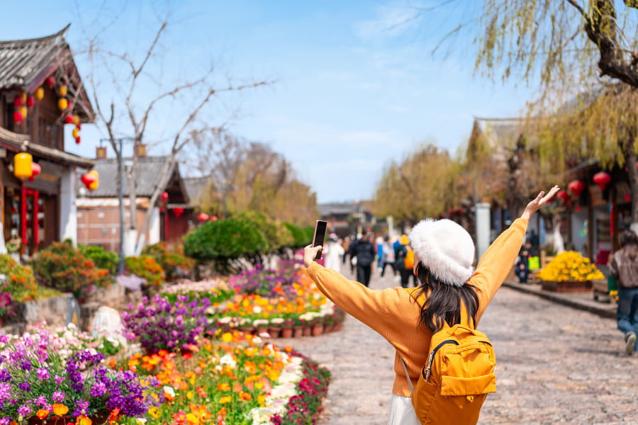 Turista caminando por una pintoresca calle tradicional de China rodeada de coloridos jardines y arquitectura típica, mientras toma una fotografía con su teléfono móvil. La imagen representa el turismo, los viajes y la cultura china en un destino histórico.
