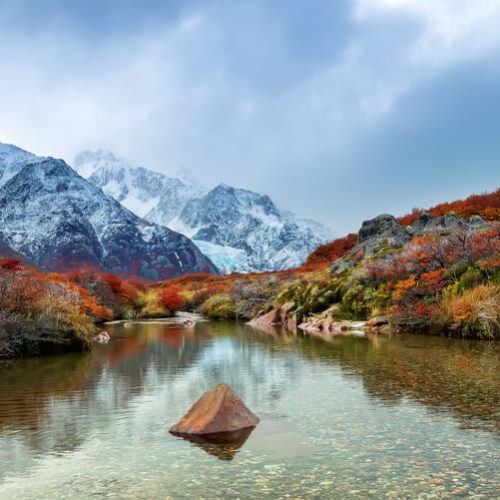 Esta imagen muestra un paisaje natural de alta montaña, caracterizado por el contraste entre los colores otoñales de la vegetación baja y la frialdad de las cumbres nevadas.