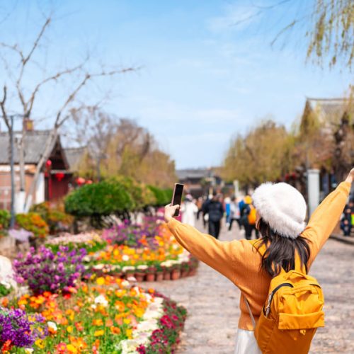 Viajera de espaldas disfrutando y fotografiando un antiguo pueblo chino con arquitectura tradicional y jardines de flores en un día soleado.